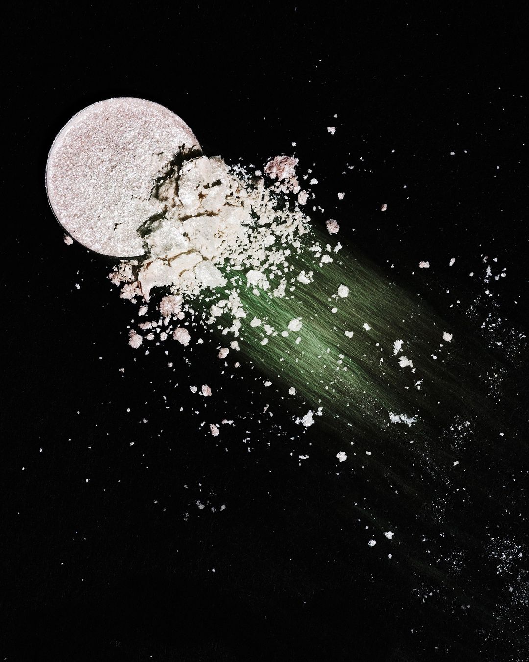 Close-up shot of a round eye shadow palette with shattered powder in iridescent shades, set against a stark black background.