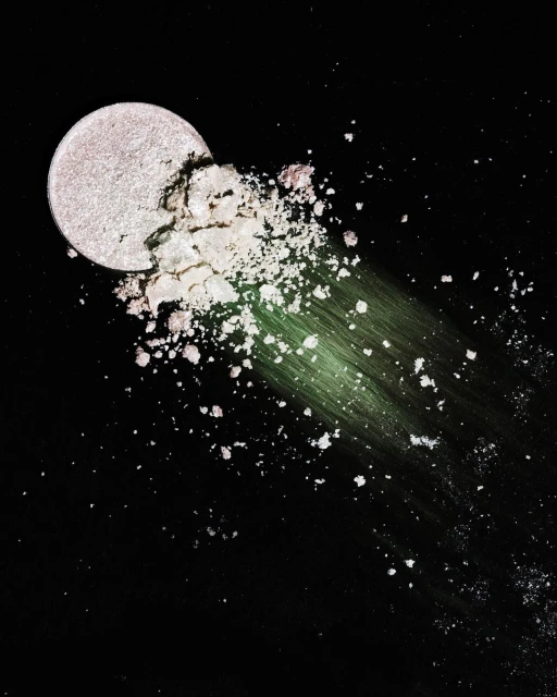 Close-up shot of a round eye shadow palette with shattered powder in iridescent shades, set against a stark black background.