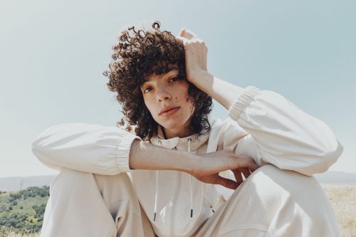 Portrait of a curly-haired woman in a white track suit, sitting outdoors against a clear sky and grassy hills.