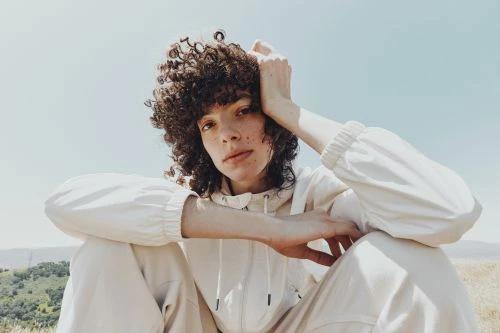 Portrait of a curly-haired woman in a white track suit, sitting outdoors against a clear sky and grassy hills.