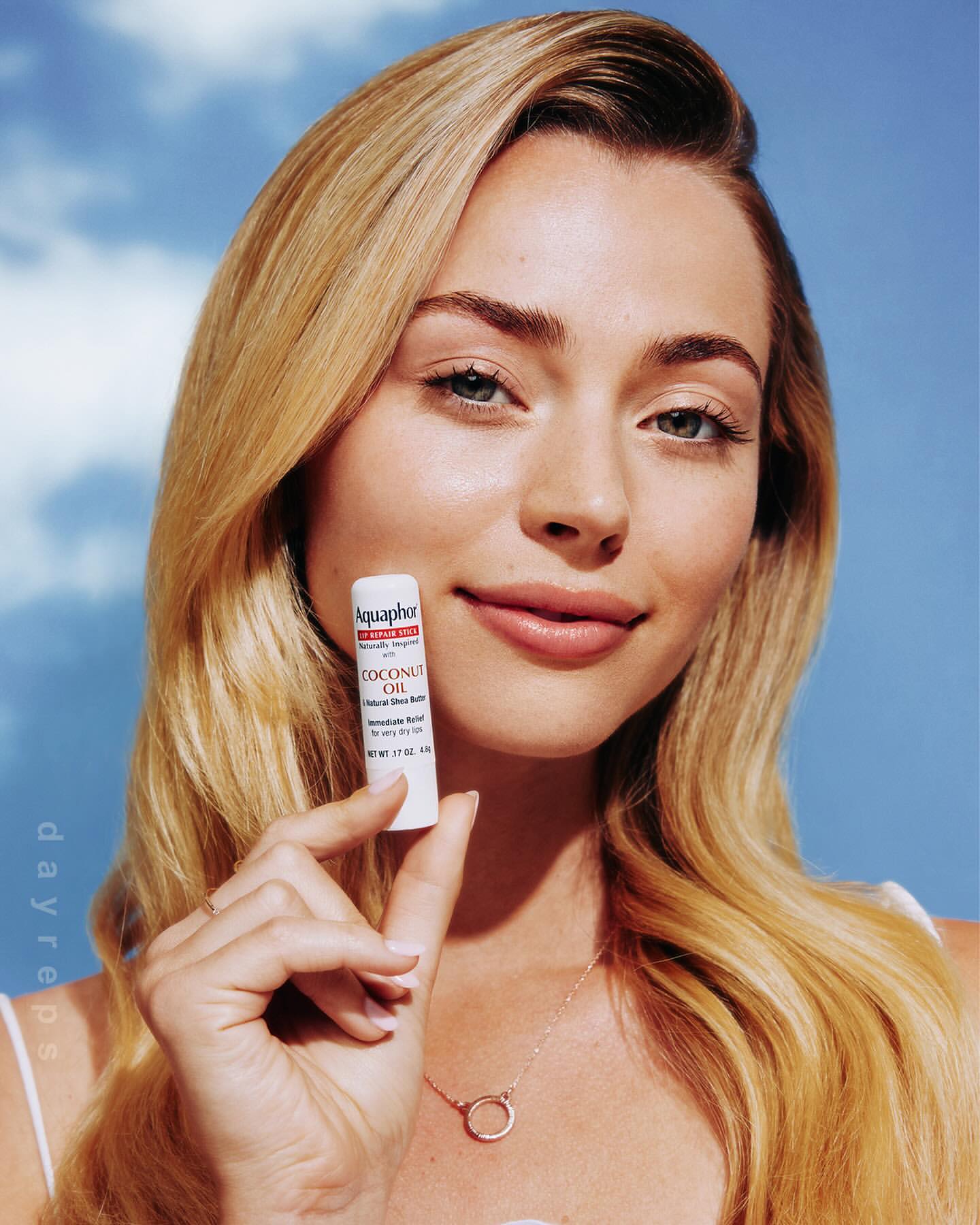 A blonde woman smiles while holding Aquaphor Coconut Oil lip balm in an eye-level medium shot, with a blue sky background.