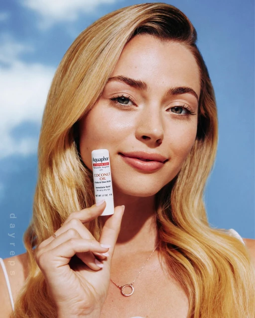 A blonde woman smiles while holding Aquaphor Coconut Oil lip balm in an eye-level medium shot, with a blue sky background.