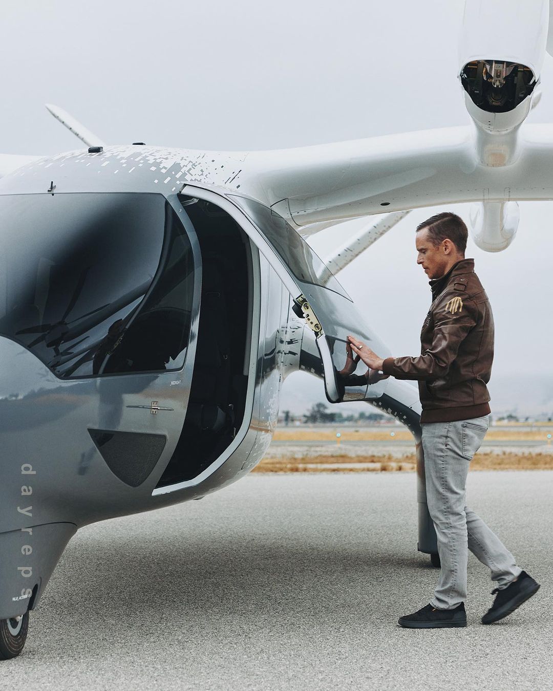 Adult man in a brown jacket standing on an airstrip, opening the door of a gray and white passenger drone or electric vertical takeoff and landing aircraft.