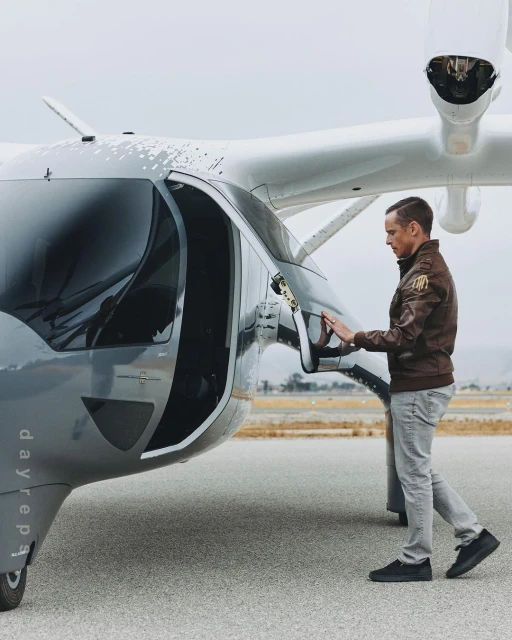 Adult man in a brown jacket standing on an airstrip, opening the door of a gray and white passenger drone or electric vertical takeoff and landing aircraft.