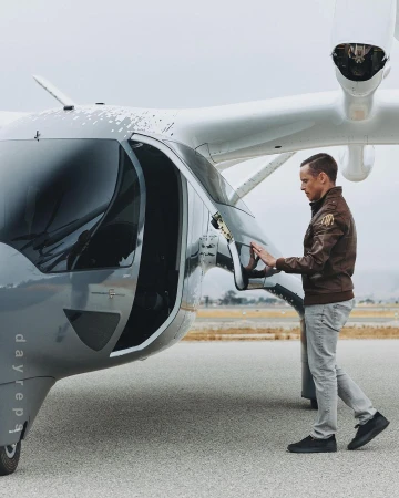 Adult man in a brown jacket standing on an airstrip, opening the door of a gray and white passenger drone or electric vertical takeoff and landing aircraft.