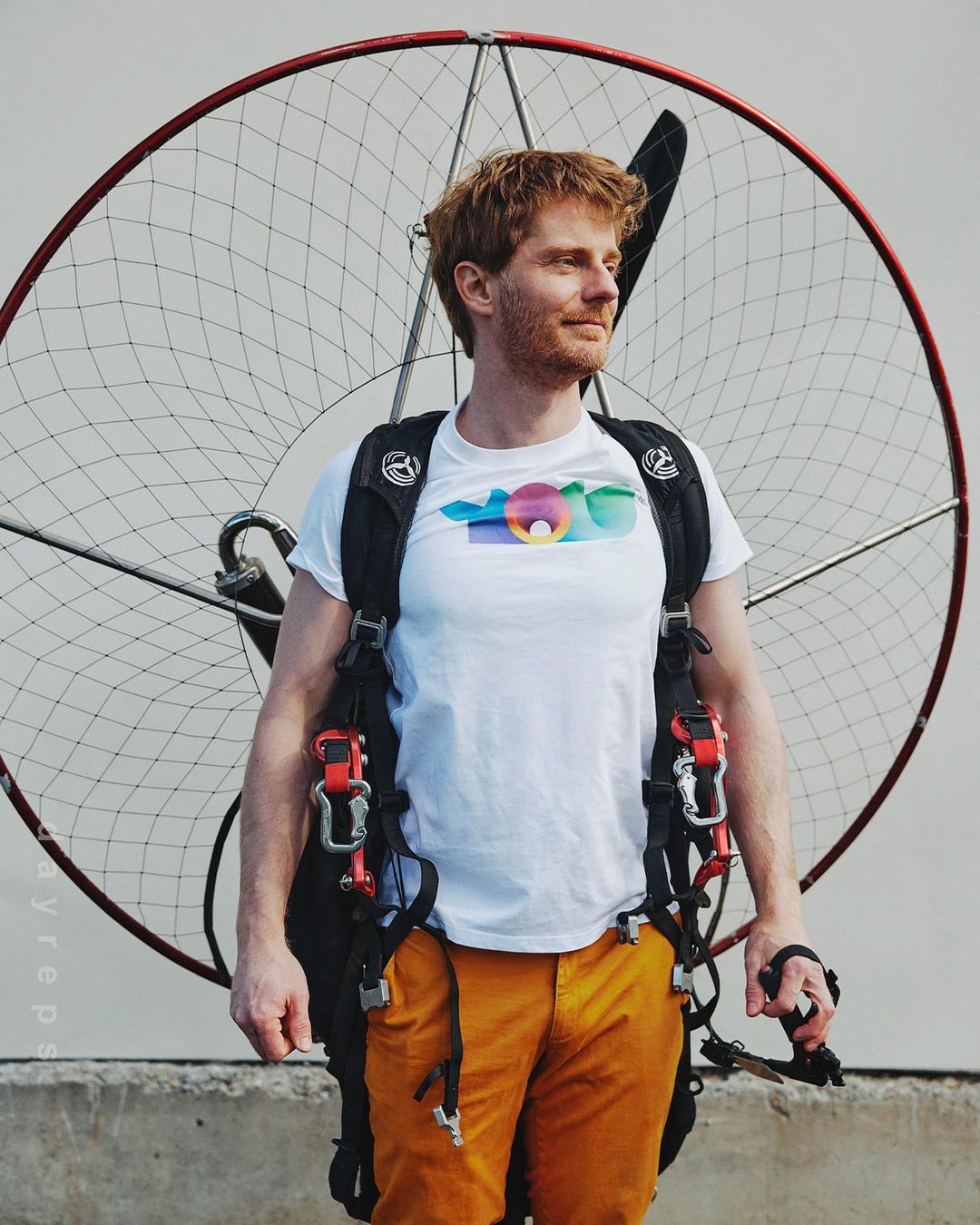 Man with red hair wearing a white t-shirt and orange pants standing next to a motorized paraglider on an overcast day.