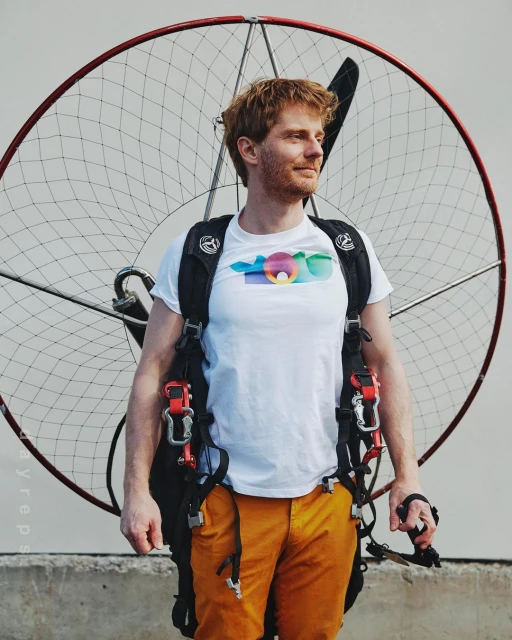 Man with red hair wearing a white t-shirt and orange pants standing next to a motorized paraglider on an overcast day.