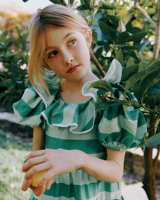 Young girl with blonde hair and heterochromia poses outdoors in green and white striped dress holding a lemon, surrounded by green foliage.