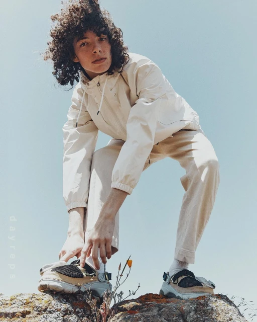 Stylish individual crouches on a rock wearing beige Crocs, matching tracksuit, set against a bright blue sky backdrop in an outdoor setting.