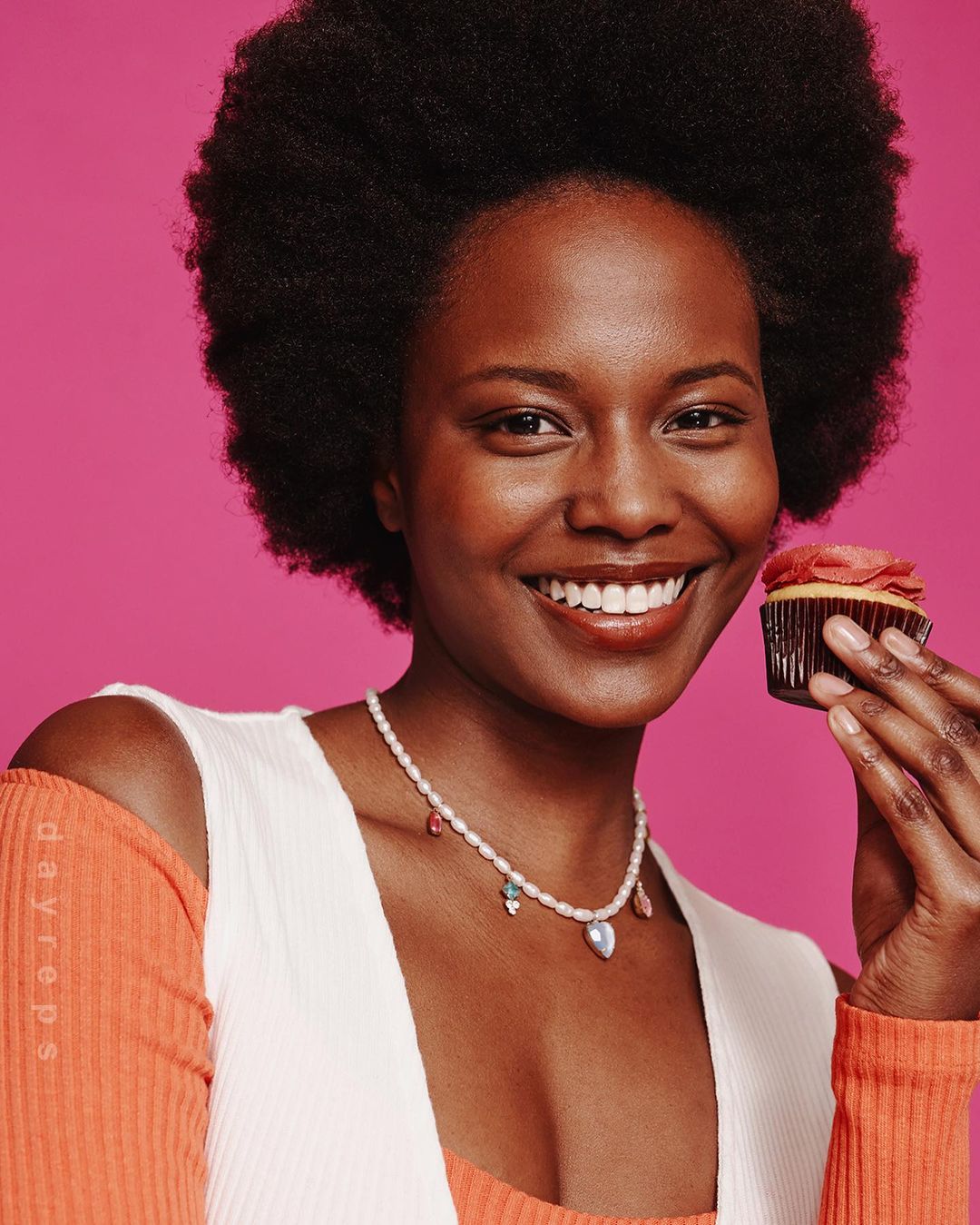 Smiling African American woman holds a pink frosted cupcake against a bright pink background, wearing a pearl necklace and orange top.