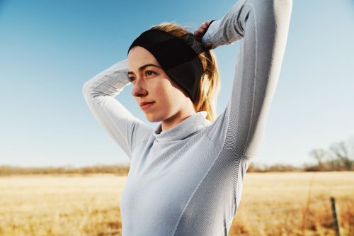 Woman in athletic wear adjusts her black headband outdoors in a rural setting with a blue sky and a field of dry grass.