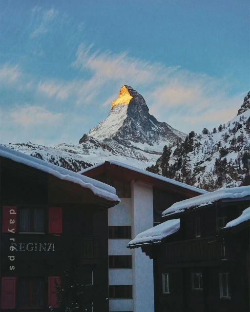 Mountain peak covered in snow is visible above buildings with snow-covered roofs under a blue and cloudy sky in an outdoor winter scene.