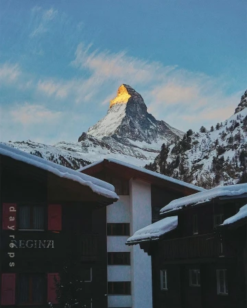 Mountain peak covered in snow is visible above buildings with snow-covered roofs under a blue and cloudy sky in an outdoor winter scene.
