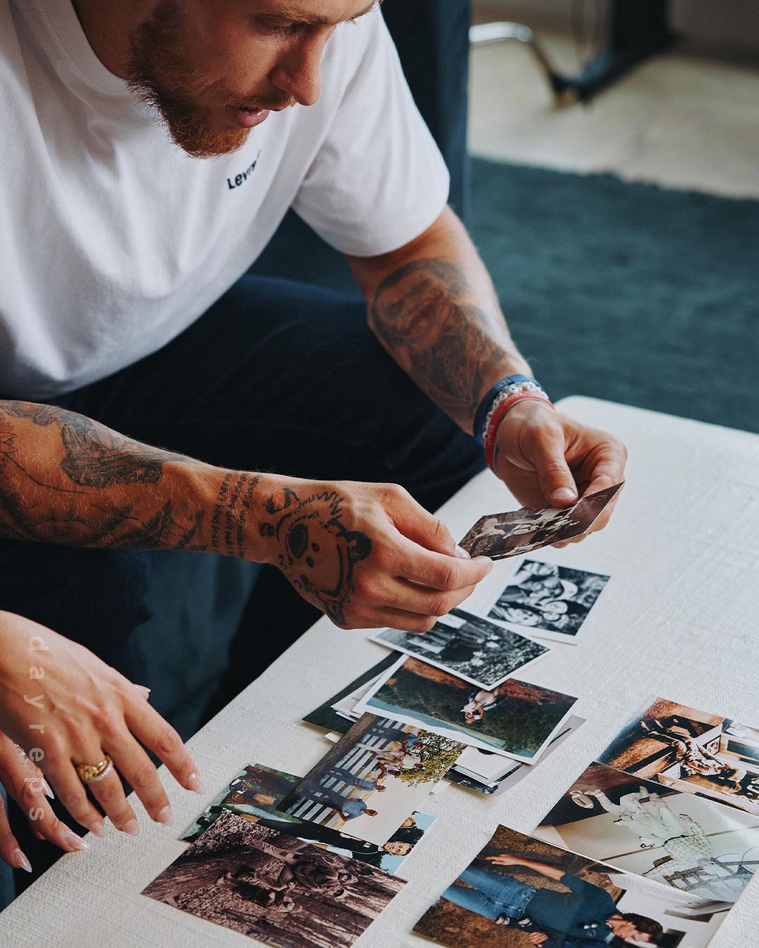 Man looking at photographs on a white table, with a woman's hand visible. He is wearing a white shirt with tattoos on his arms.