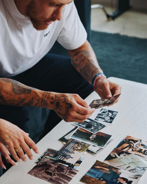Man looking at photographs on a white table, with a woman's hand visible. He is wearing a white shirt with tattoos on his arms.