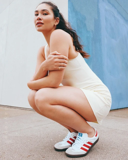 Athletic woman in a cream dress and red and white Adidas sneakers crouching outdoors against a blue and white wall.