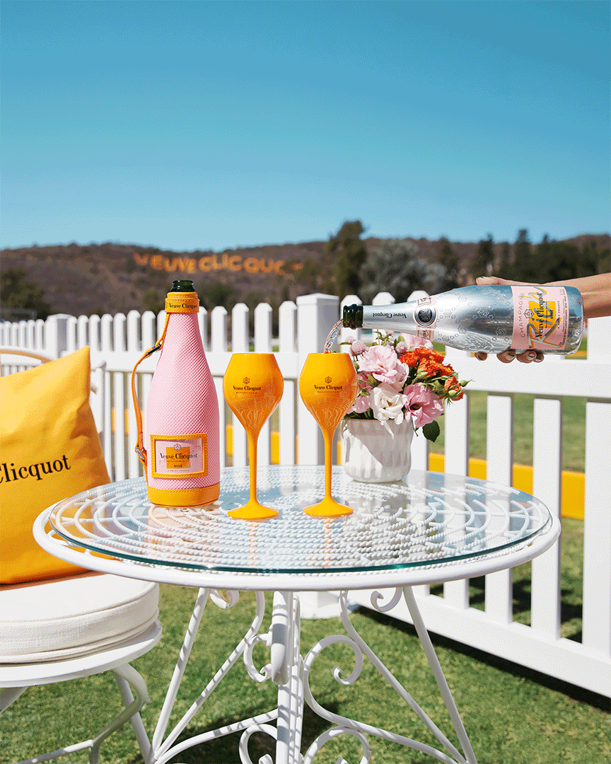 Outdoor setting with champagne: bottle with pink cover, two filled yellow flutes, a bouquet, and a pillow on a white wrought-iron table.