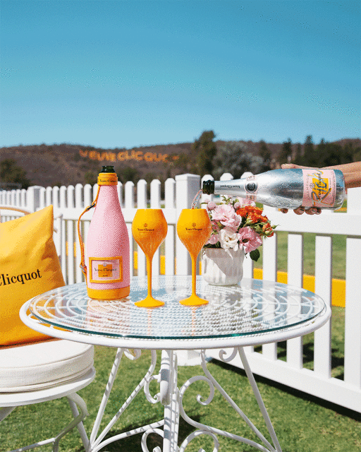 Outdoor setting with champagne: bottle with pink cover, two filled yellow flutes, a bouquet, and a pillow on a white wrought-iron table.