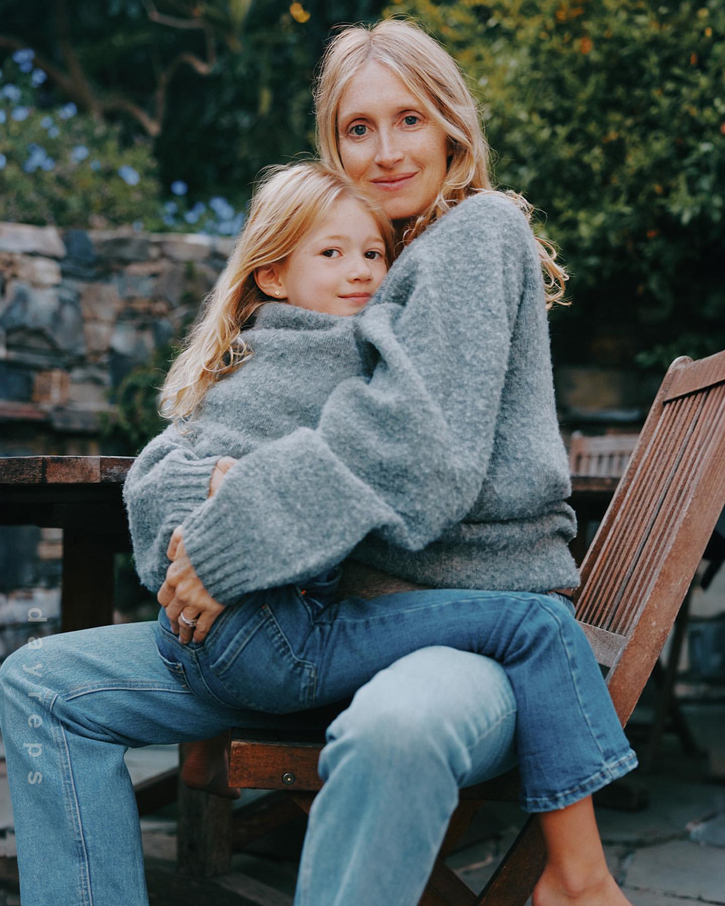 A mother and child embrace outdoors in matching grey sweaters and blue jeans, seated on a wooden bench surrounded by stone and greenery.