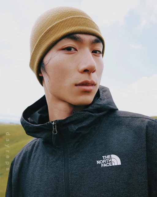 Portrait of an Asian man outdoors wearing a beanie and hooded jacket with the North Face logo, standing against a sky and grassy background.