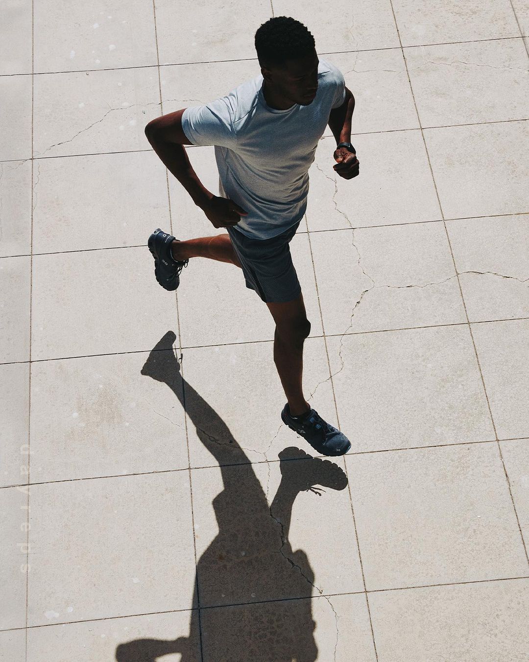 Aerial view of Black male athlete running in grey athletic wear on a tiled surface with his shadow visible on a sunny day.