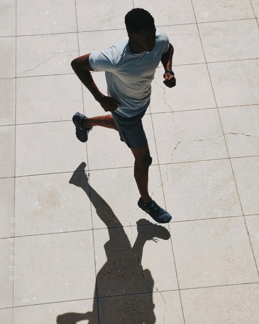 Aerial view of Black male athlete running in grey athletic wear on a tiled surface with his shadow visible on a sunny day.