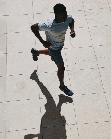 Aerial view of Black male athlete running in grey athletic wear on a tiled surface with his shadow visible on a sunny day.