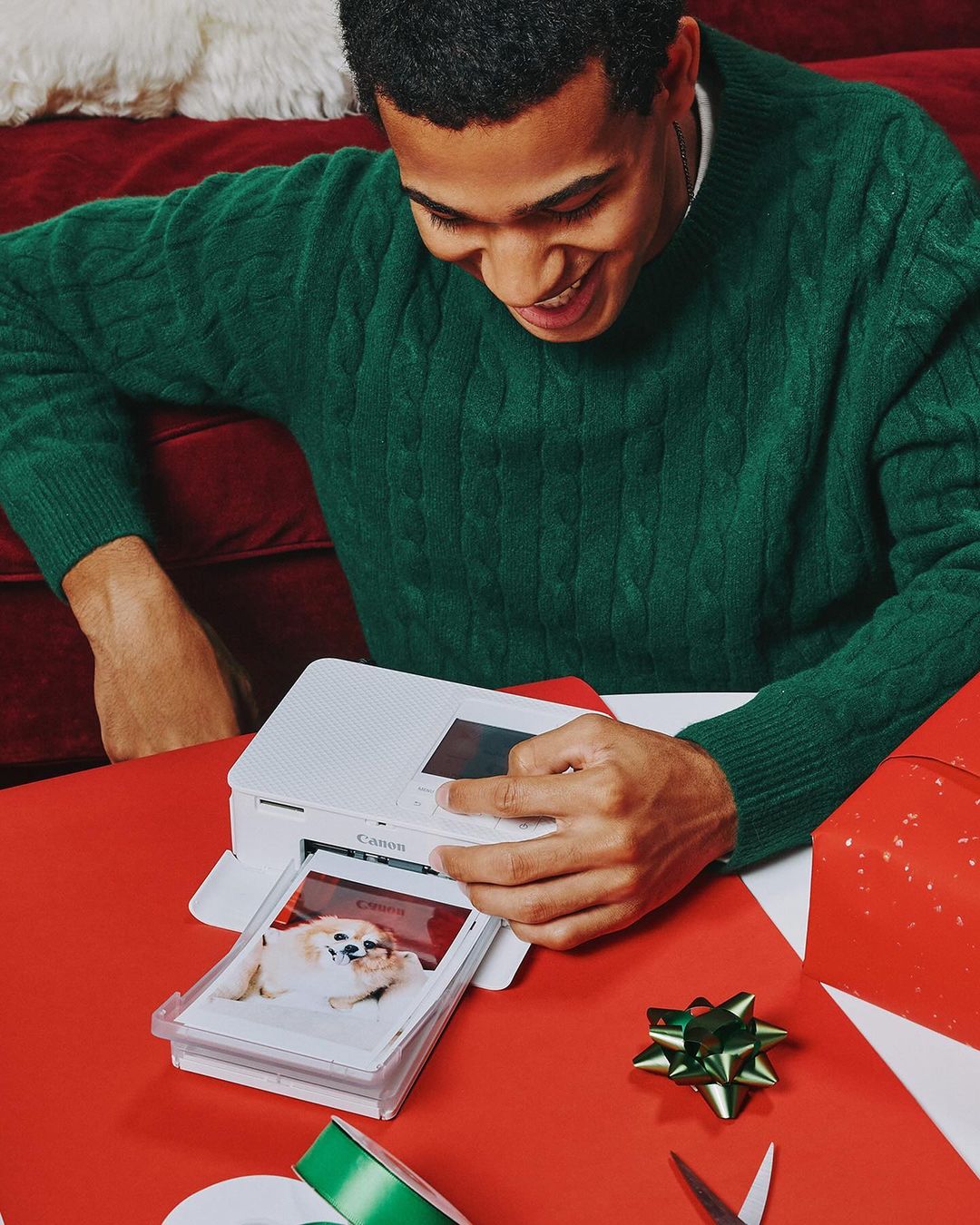 Smiling man in a green sweater prints a photo of a dog using a compact photo printer on a red table with craft supplies.