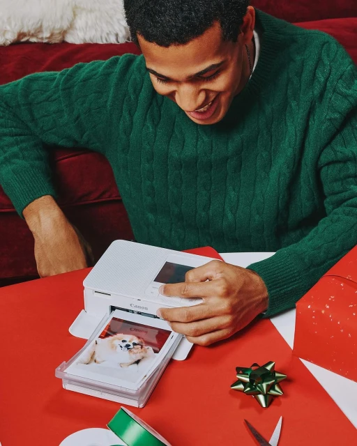 Smiling man in a green sweater prints a photo of a dog using a compact photo printer on a red table with craft supplies.