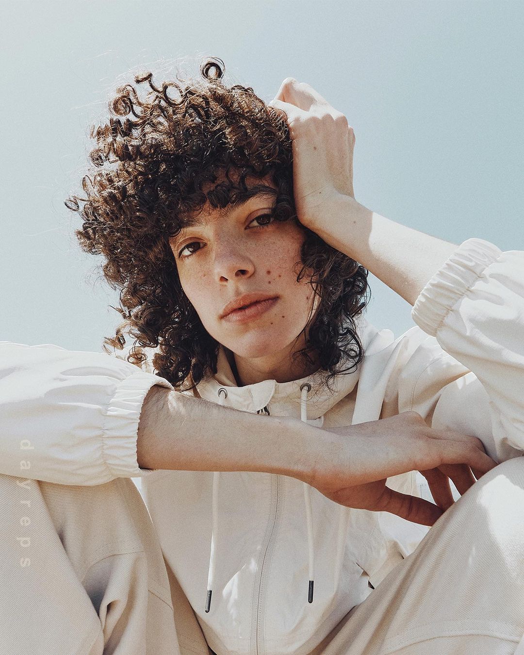Portrait of a curly-haired adult posing in an off-white hooded jacket and pants against a clear sky background on a sunny day.