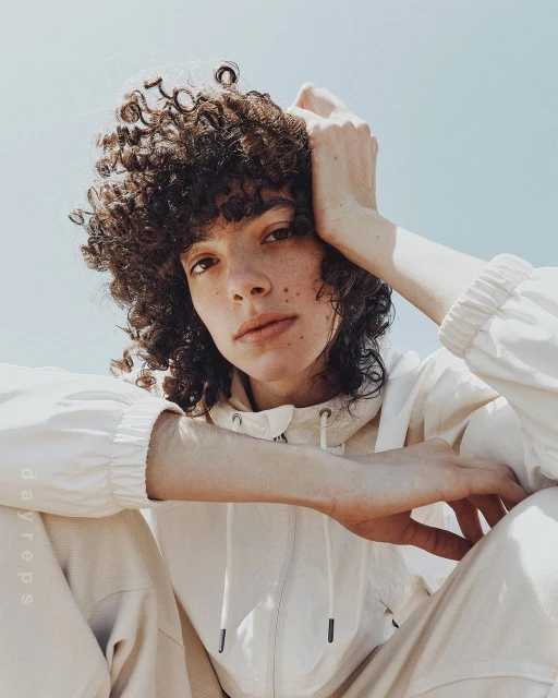 Portrait of a curly-haired adult posing in an off-white hooded jacket and pants against a clear sky background on a sunny day.