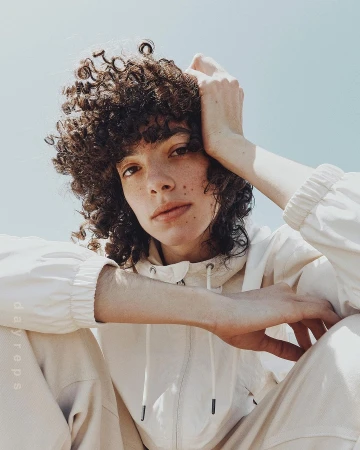 Portrait of a curly-haired adult posing in an off-white hooded jacket and pants against a clear sky background on a sunny day.