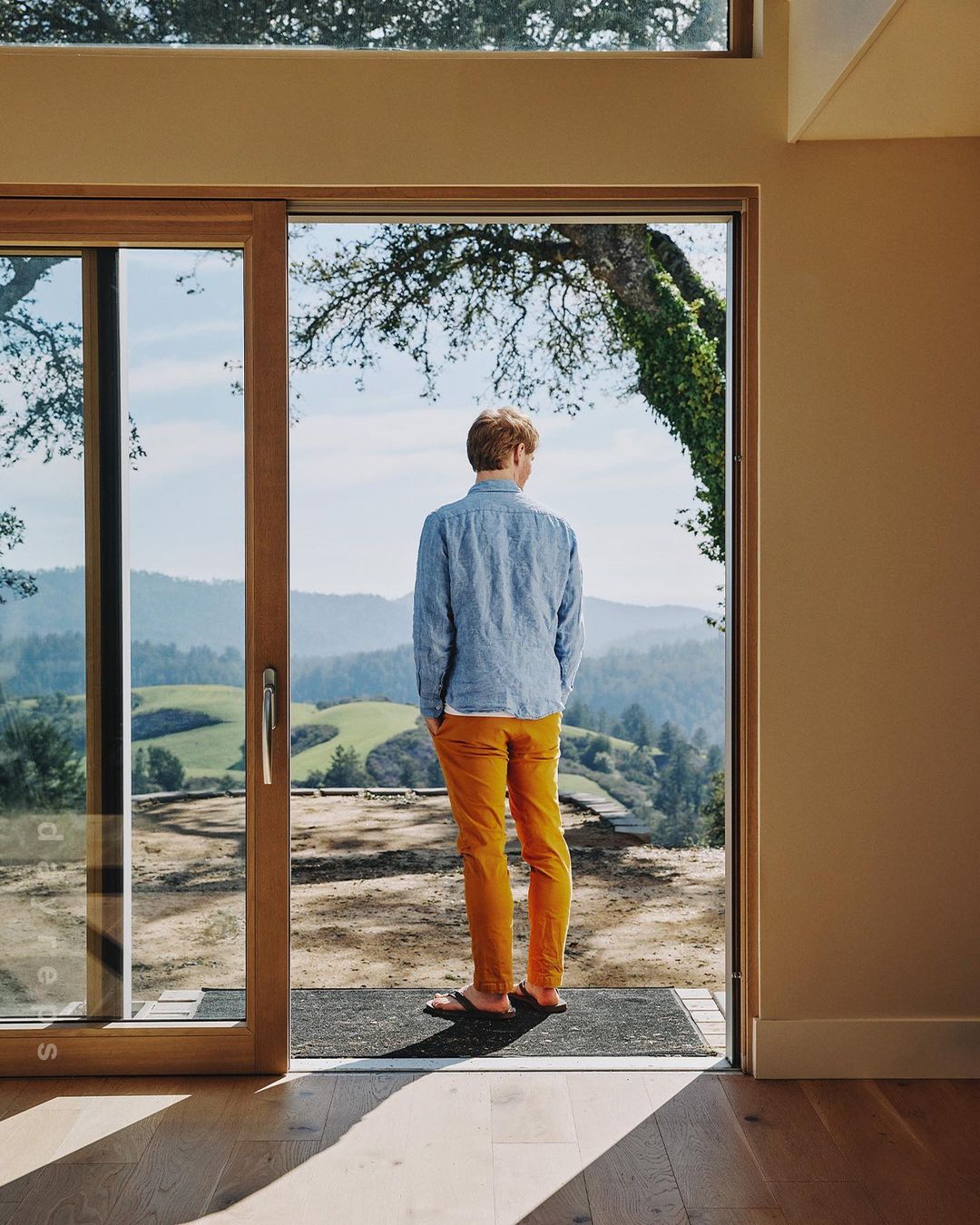 Back view of an adult male standing in the doorway looking out to a landscape of rolling hills and green fields on a sunny day.