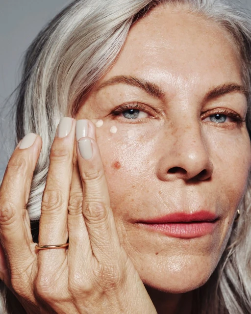 Close-up of a mature woman with gray hair applying light-toned cream under her eye. The focus is on her face, skin texture and beauty product.