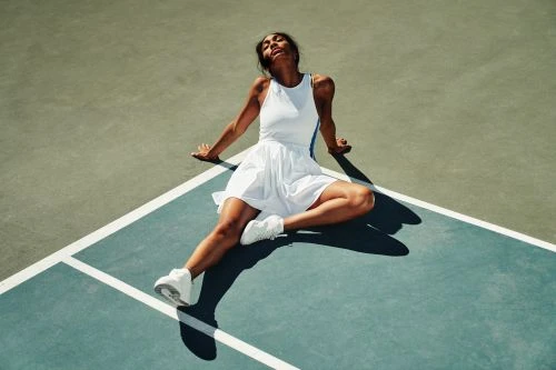 Female tennis player in white dress and sneakers sits on the court with head back in the sunshine on a blue and grey court.