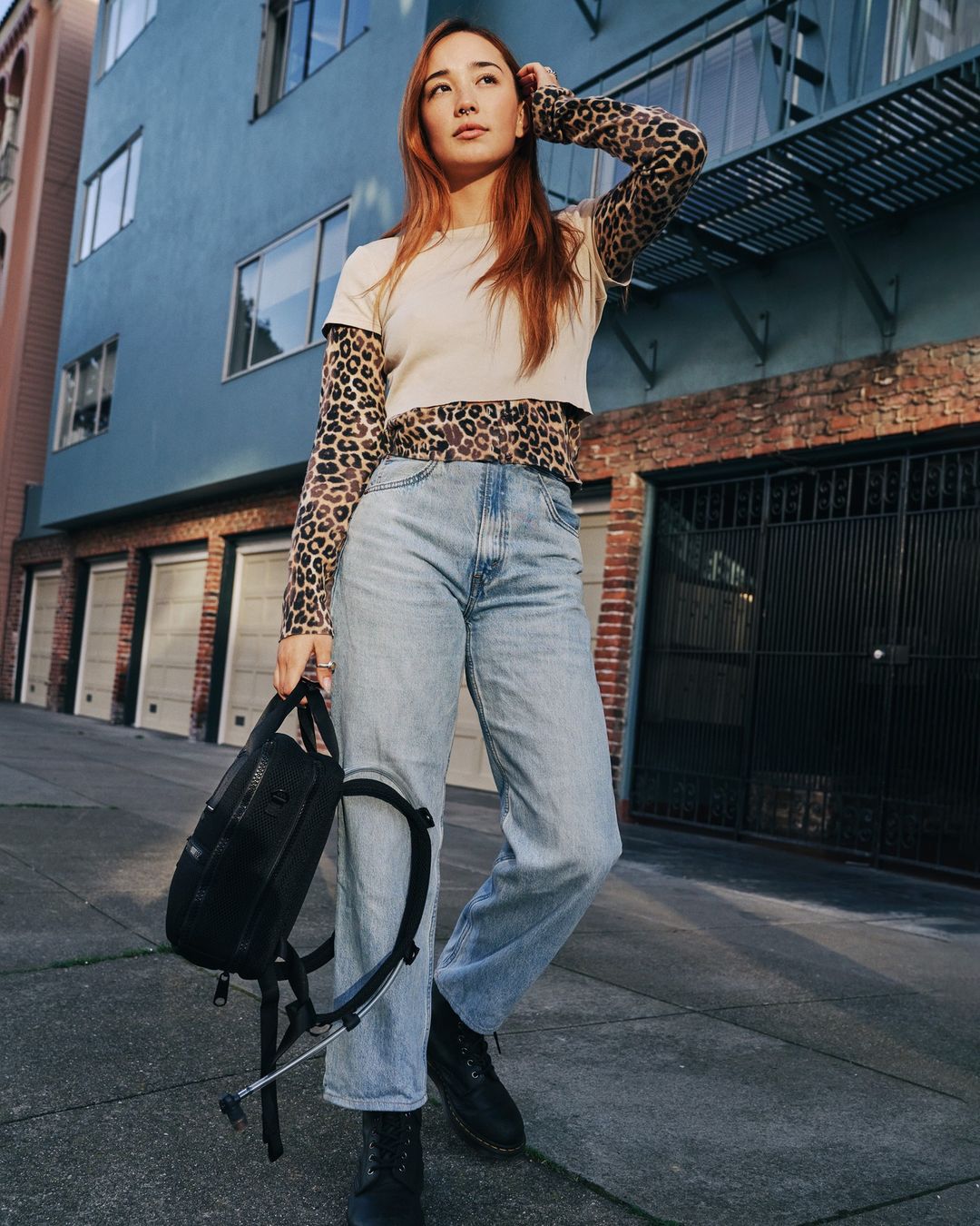 A woman with auburn hair wears a leopard print top with a neutral-colored t-shirt and blue jeans while carrying a black backpack on a city street.