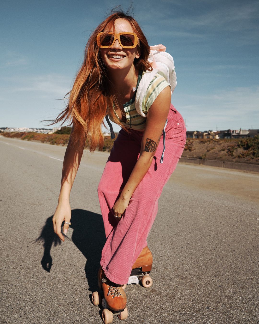 A smiling red-haired woman roller skating down an asphalt road on a sunny day, wearing pink pants and yellow sunglasses.