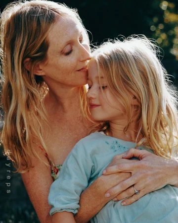 A tender moment of a fair-skinned woman with long blonde hair kissing a fair-skinned child on the forehead with a blurred background.