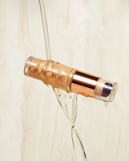 Close-up studio shot of Quickie brand cosmetic product with tan liquid dripping on it, against a beige background with light veins.