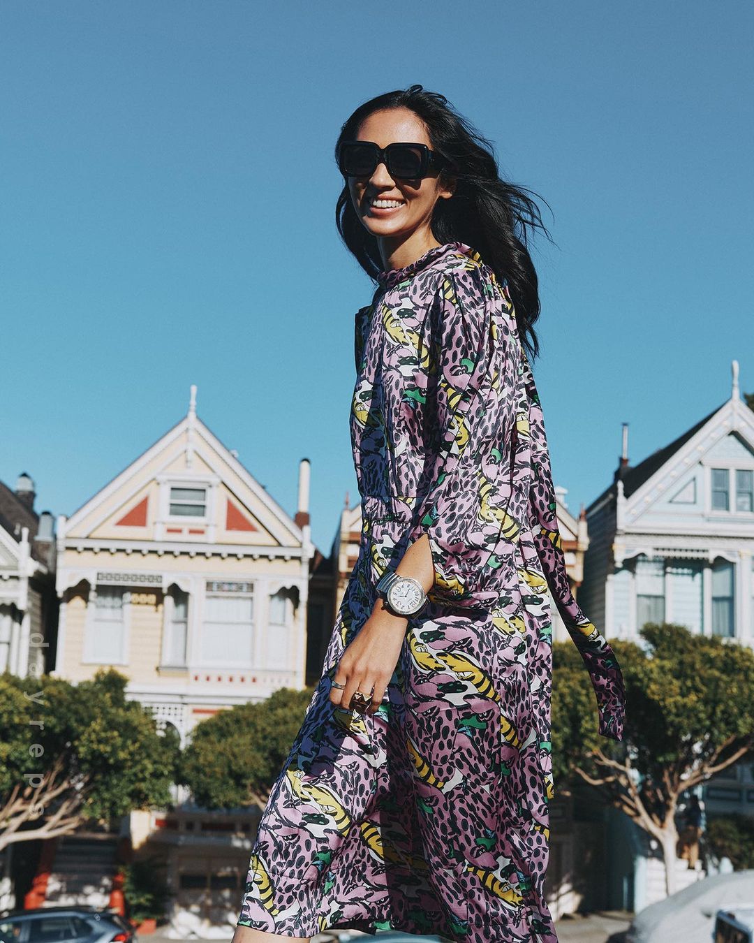 Smiling woman in printed purple maxi dress and sunglasses walks near colorful houses on a sunny day in an urban outdoor environment.