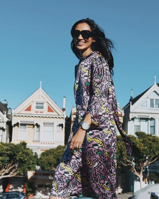 Smiling woman in printed purple maxi dress and sunglasses walks near colorful houses on a sunny day in an urban outdoor environment.