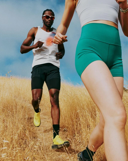 Two athletic runners on a grassy hillside: one male, one partially visible female, wearing activewear and athletic shoes. Sunny day.