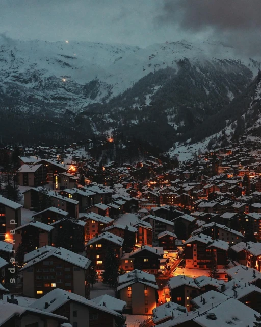 Snowy mountain village at night with lights twinkling in buildings under snow-covered mountains, creating a serene winter landscape.