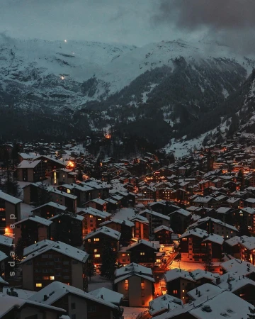 Snowy mountain village at night with lights twinkling in buildings under snow-covered mountains, creating a serene winter landscape.
