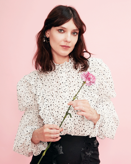 A woman with shoulder-length brown hair in a white and black patterned blouse holds a pink flower against a pink backdrop.