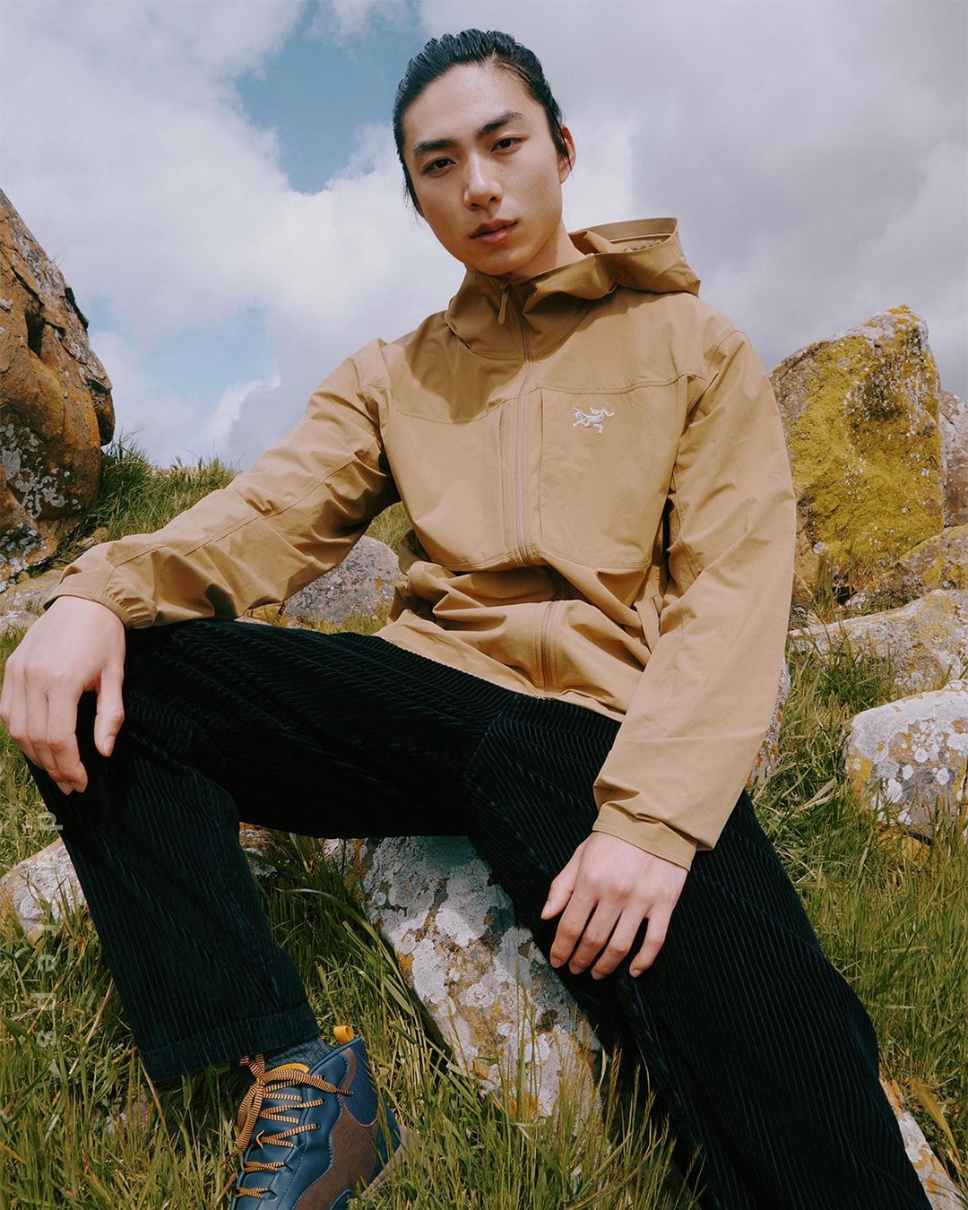 Adult male poses outdoors on rock wearing a tan hooded jacket and dark corduroy pants in a lifestyle fashion photo against a cloudy sky.