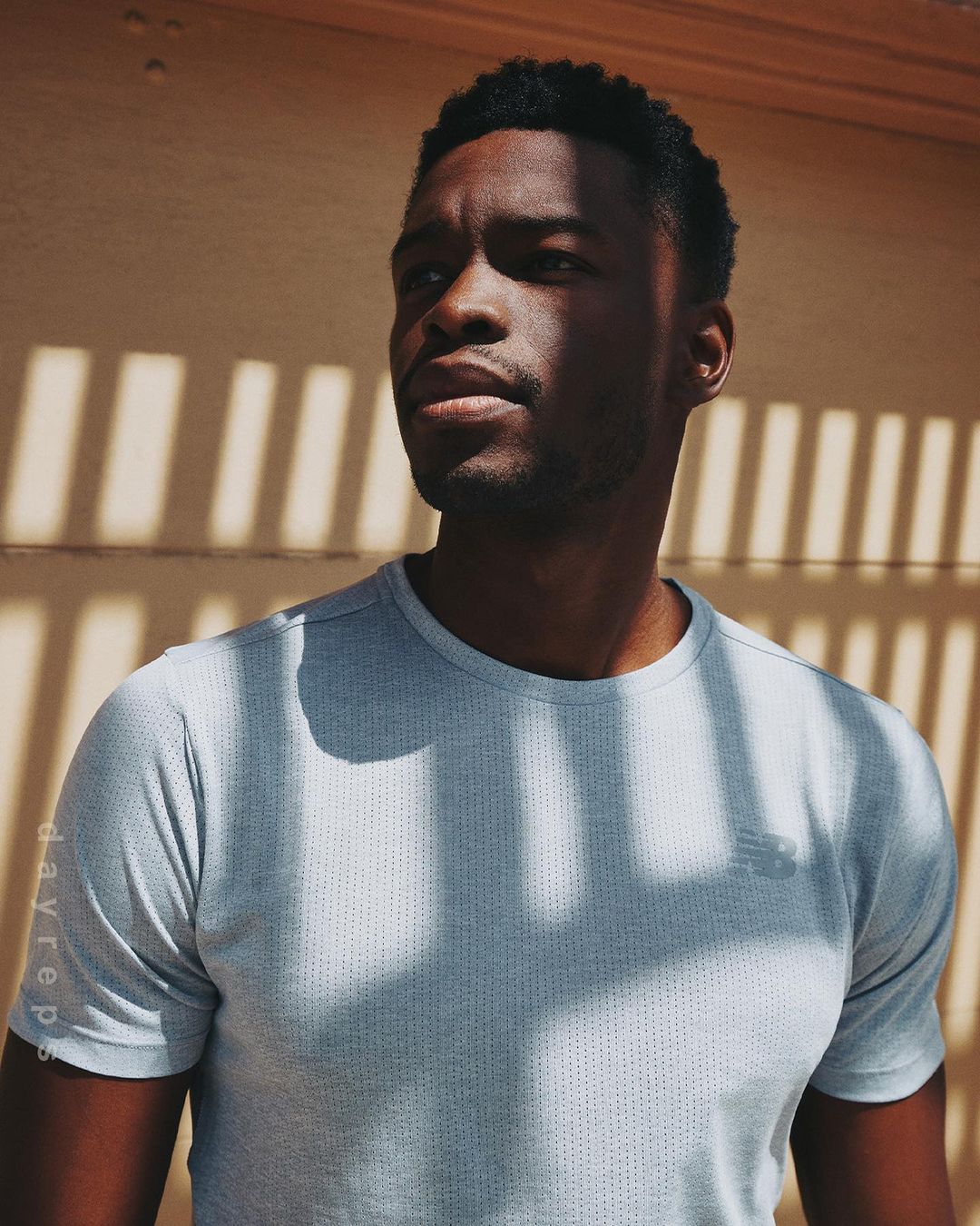 Portrait of a Black male in a grey t-shirt, standing outdoors in a bright, natural light with architectural shadow patterns.