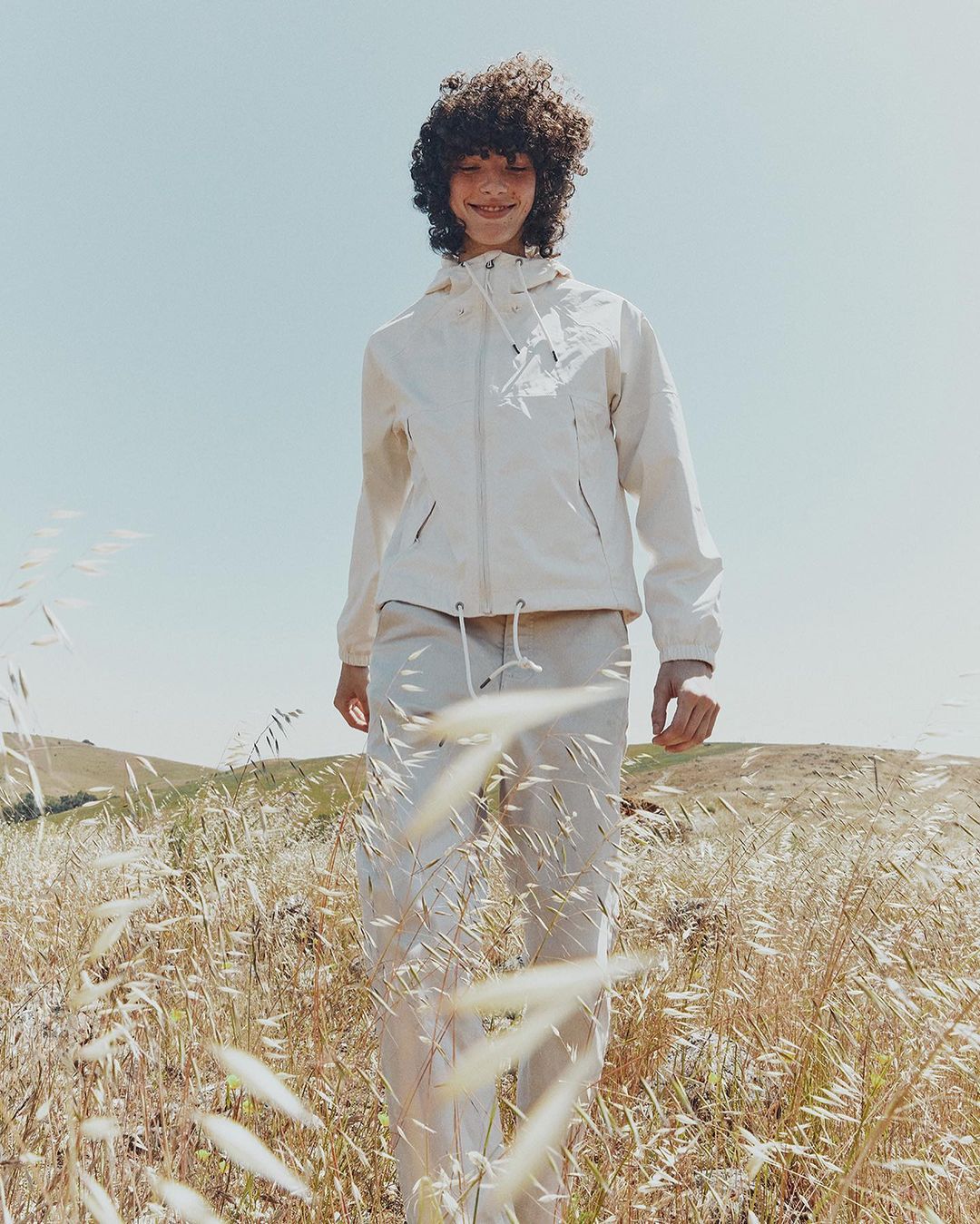 Teenager in an off-white windbreaker jacket and pants standing in a sunny, grassy field on a clear day, fashion editorial.