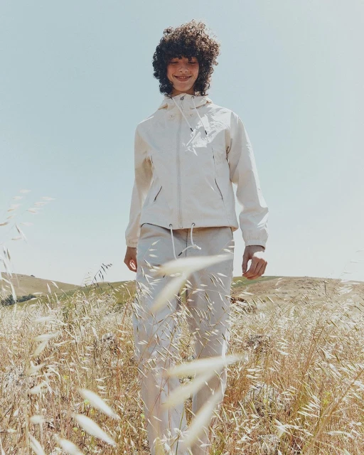 Teenager in an off-white windbreaker jacket and pants standing in a sunny, grassy field on a clear day, fashion editorial.