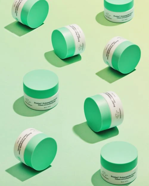 Several jars of green and white skincare cream are arranged on a light green and yellow gradient background, creating a clean and modern beauty product display.